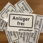 Das Bild zeigt einen Stapel von Aufklebern mit der Aufschrift „Anlüger frei“ in einem stilisierten Verkehrszeichendesign. Das Bild zeigt mehrere rechteckige Aufkleber, die übereinanderliegen und einen kleinen Stapel bilden. Die Aufkleber sind weiß mit schwarzem Rand und schwarzer Schrift. Auf jedem Aufkleber steht in großen, deutlichen Buchstaben „Anlüger frei“. Unter dem Text ist in kleinerer Schrift „BERLIN CYCLING DIARY“ zu lesen. Die Aufkleber haben das Aussehen von vereinfachten Verkehrszeichen. Der Stapel liegt auf einer braunen, hölzernen Oberfläche, die wie ein Tisch oder eine Arbeitsplatte aussieht. Die Beleuchtung ist neutral und die Gesamtstimmung des Bildes ist sachlich und unaufgeregt. Die Fokussierung liegt auf den Aufklebern, der Hintergrund ist unscharf. Es gibt keine Personen oder Tiere auf dem Bild.